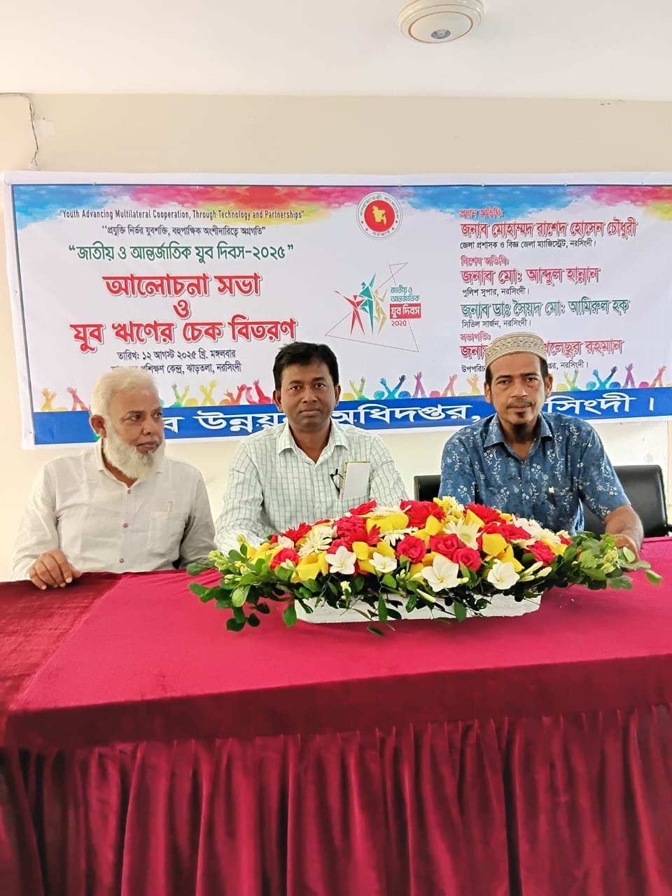 Indoor seminar; two men at a table with flowers.